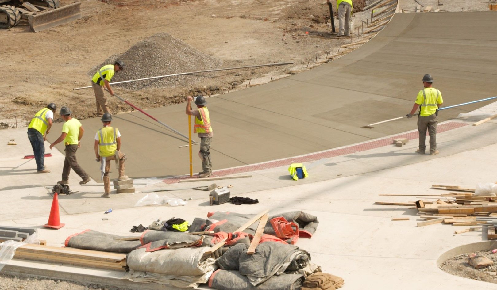 A crew in hard hats and high-visibility vests screeding and floating a freshly poured concrete slab on a construction site, lumber and pipe stacked along the finished edge