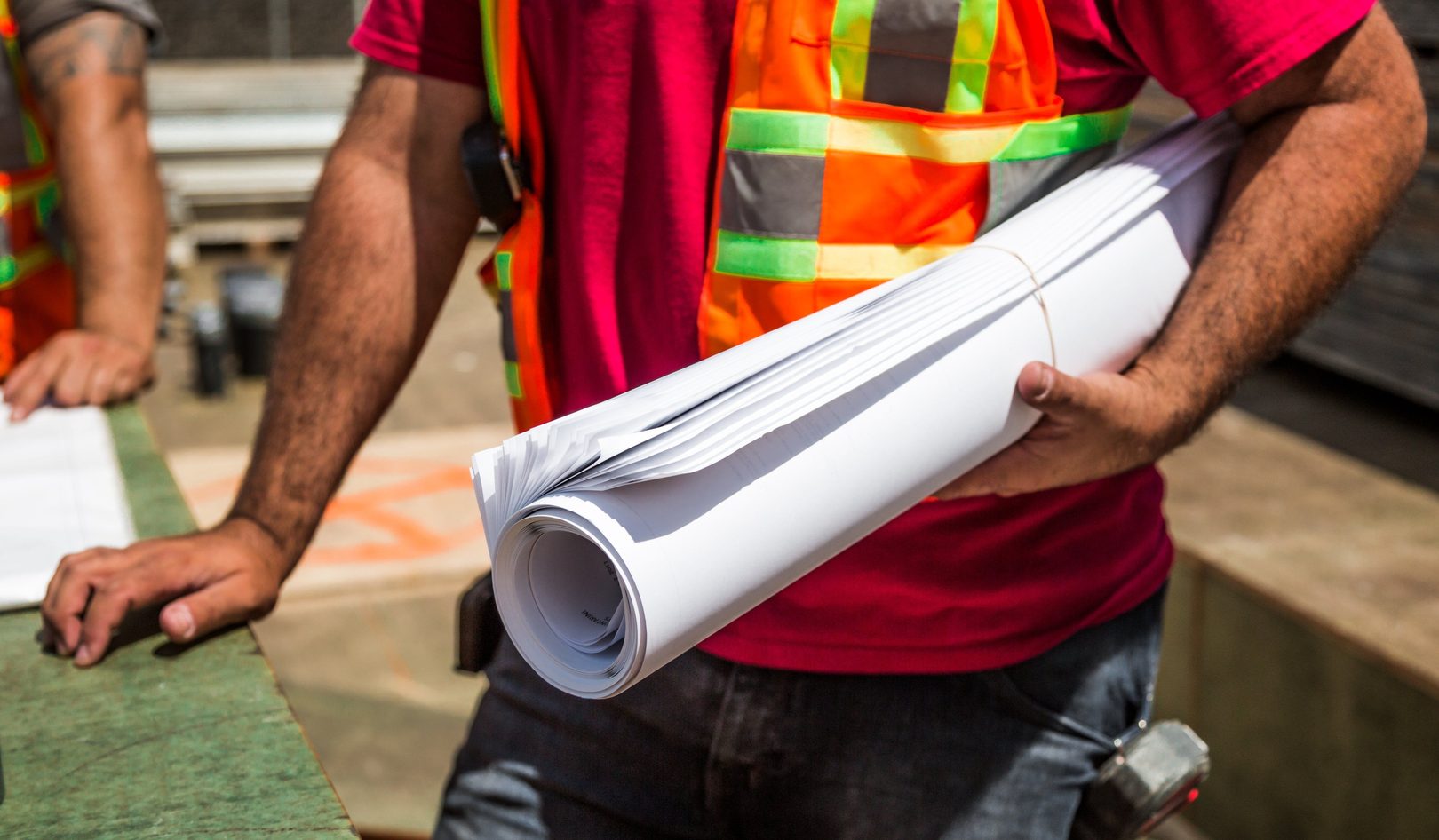 A construction worker in a high-visibility vest carrying a rolled set of drawings under one arm on a jobsite, a second worker’s hands flat on an open drawing sheet on the plywood table beside him