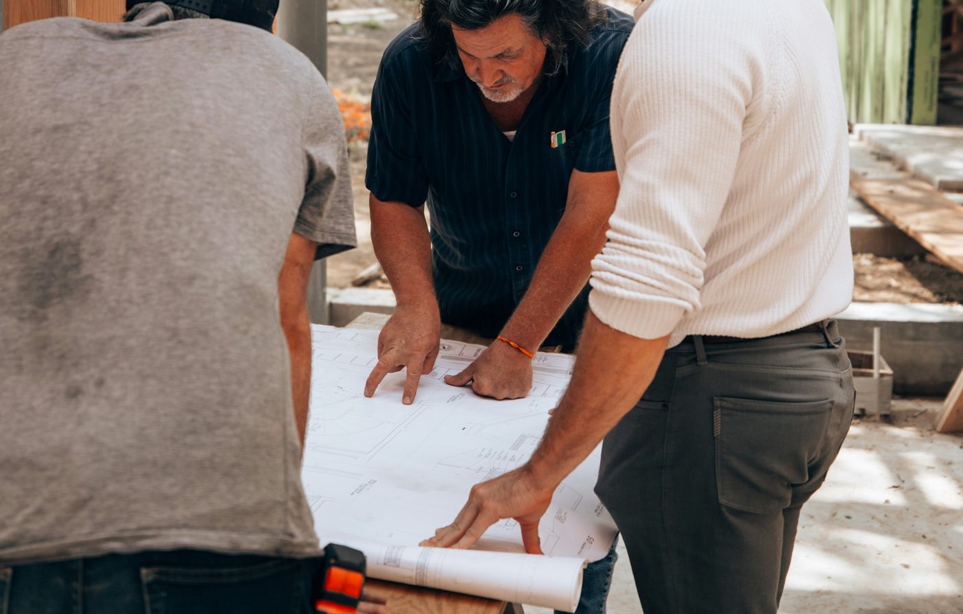 Three people leaning over a large paper drawing set spread on a plywood table at an active jobsite, hands pointing at the sheet, a second roll of drawings still rolled up beside it