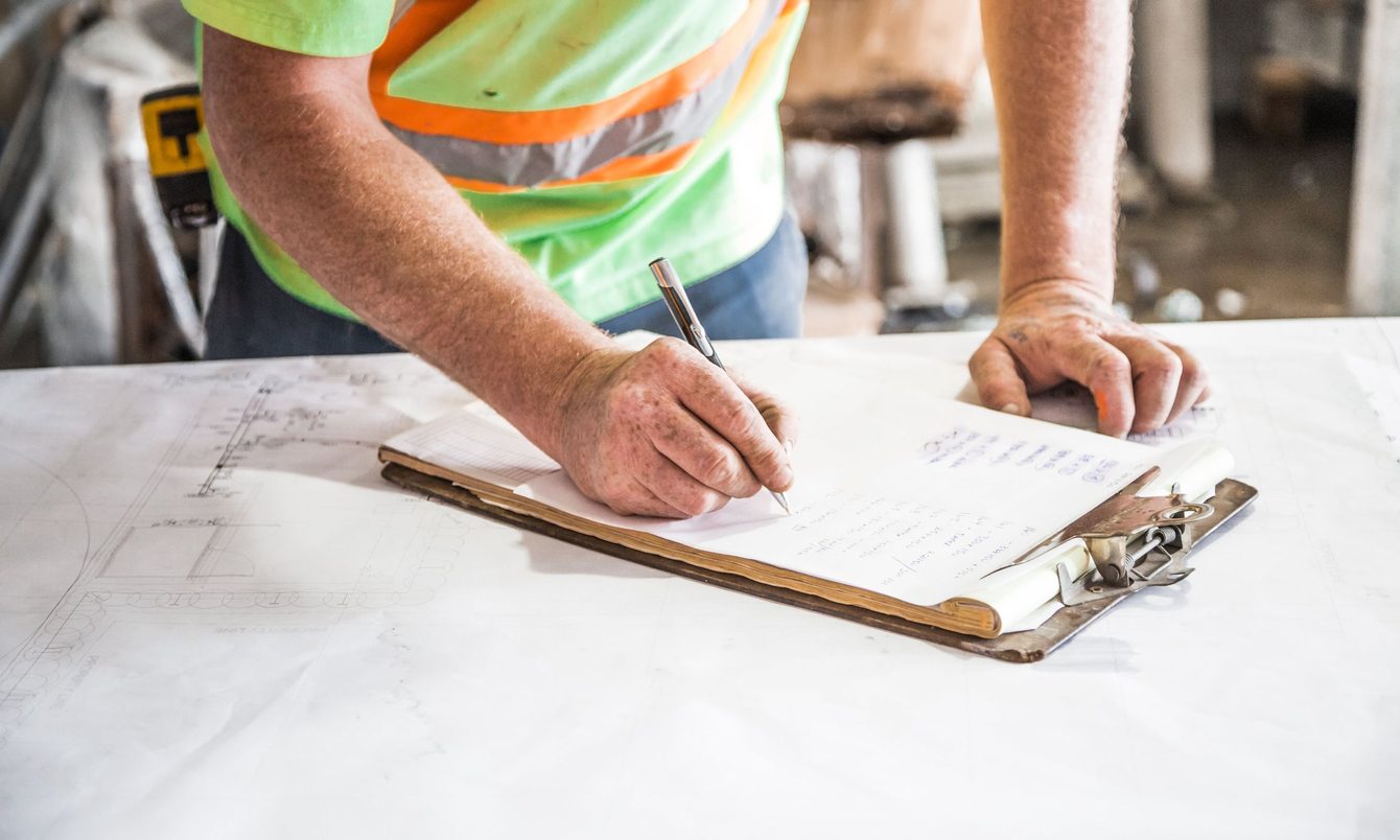 A worker in a hi-vis vest writing on a clipboard resting on an open drawing at a construction site, with a tape measure beside it